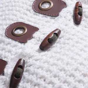 Nahaufnahme eines weißen Strickpullovers mit braunen Lederflicken und Holz-Knöpfen.