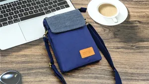 Blue canvas mini crossbody phone bag detail view on wooden table with laptop and coffee.