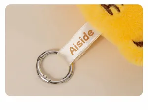 Plush toy keychain detail shot featuring soft yellow fabric, branded ribbon tag and metal ring.