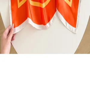 Detail foulard en soie orange avec motif geometrique jaune et main tenant le coin du tissu.