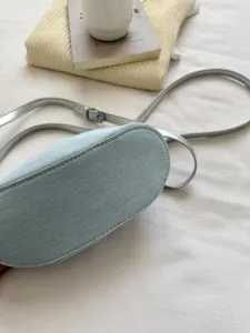 Detail shot of light blue denim crossbody bag with adjustable silver strap on white bedding.