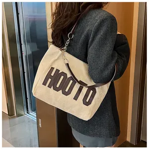 Detail shot of beige tote bag with brown HOOTO letters and chain strap worn by woman.