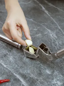 Stainless steel garlic press detail showing hand placing garlic cloves on dark stone surface.