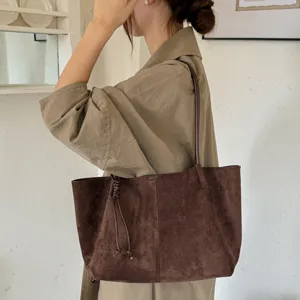 Close-up detail of a brown suede shoulder tote bag worn by a model in a minimalist room.