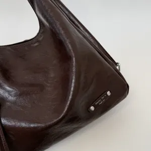 Dark brown leather shoulder bag close-up showing metallic label and soft material texture.