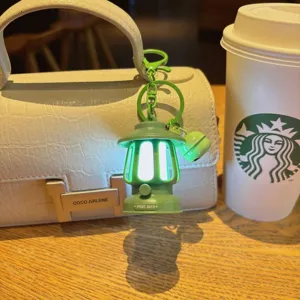 Close-up view of a mini green camping lantern keychain glowing, hanging on a textured white bag.