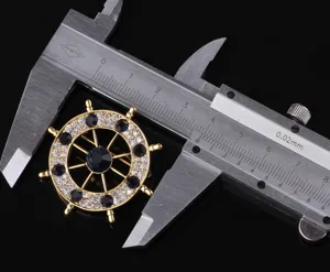 Dettaglio spilla timone nave dorata con strass e calibro per misurazione precisa.