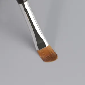 Brocha de maquillaje individual con cerdas sintéticas suaves y virola plateada en fondo gris.