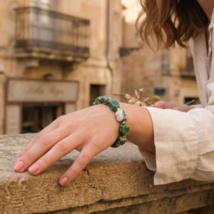 Close-up detail of irregular green stone block bracelet with gold beads worn on wrist outdoors.