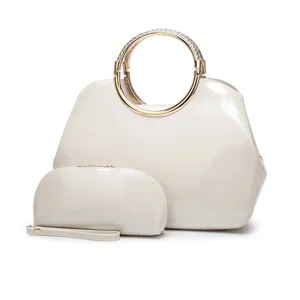 Vista detallada del conjunto de bolso de charol crema con asa redonda de metal y estuche.