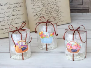Scatole regalo trasparenti cilindriche con nastri marroni e adesivi di cesti di fiori per feste.