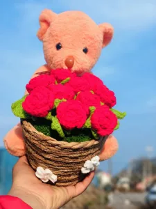 Handcrafted crochet teddy bear holding red rose bouquet in woven basket, exquisite gift detail view.
