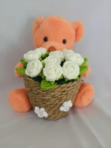 Closeup detail of handmade crochet teddy bear holding woven basket with white roses bouquet ornament.