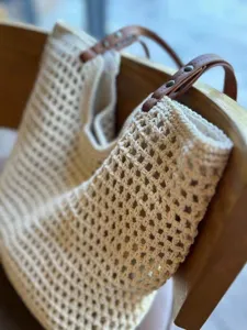 Close-up detail of beige crochet mesh tote bag with brown leather handles hanging on a chair.