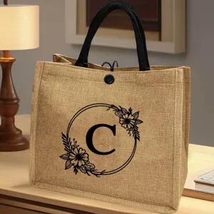 Jute-Tragetasche Detailansicht mit elegantem Blumen-Buchstaben C Druck und robusten schwarzen Griffen.