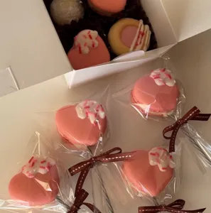Pink heart-shaped macaron lollipops in clear packaging bags with brown ribbons and a gift box.