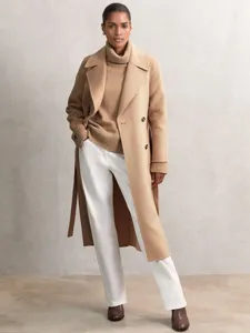 Model wearing camel turtleneck sweater and wool coat with white trousers in studio scene setup.