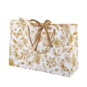 Witte papieren geschenktas met gouden bloemenprint, vlinderstrik en handige handgrepen.