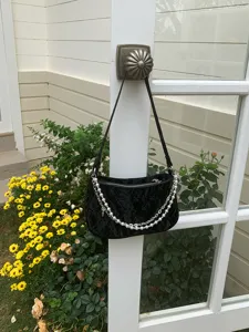Close-up of black quilted underarm bag with pearl chain hanging on white door in garden.
