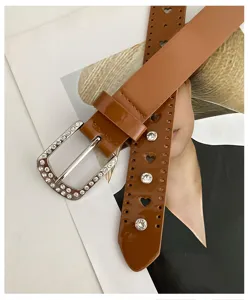 Détail de ceinture en cuir vernis marron, boucle strass, découpes cœur et accents de cristaux.