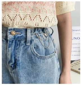 Close-up detail of stylish metal chain safety pin with heart pendant on blue denim jeans waistband.