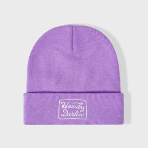 Paarse gebreide muts met witte geborduurde tekstpatch op de omslag, warme winterhoofddeksel.
