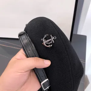Close-up detail of black wool beret with silver anchor charm and leather buckle strap.