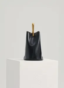 Minimalist black leather bucket bag detail featuring a striking vertical gold metal handle design.