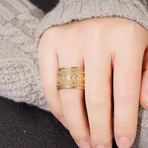 Primer plano de un anillo dorado ancho y elaborado con detalles texturados y pequeñas piedras.
