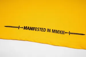 Yellow streetwear t-shirt fabric detail with black letter print and sword graphic element.