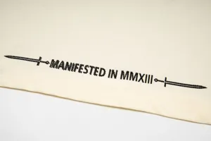 Off-white cotton fabric detail with black dagger graphic and gothic lettering for streetwear.