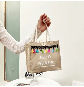 Gros plan du sac cabas en lin avec des pompons colorés, un bouton blanc et des lettres.