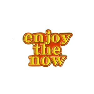 Close-up detail of the retro yellow and orange enamel pin featuring 'enjoy the now' lettering.