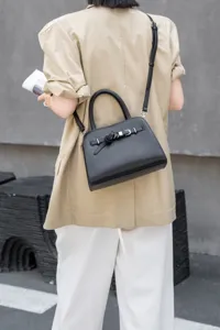 Detail shot of black leather handbag with metal buckle and shoulder strap in street setting.