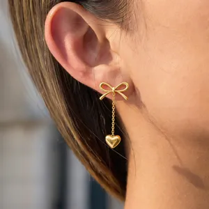 Close-up detail of stainless steel gold bow and heart drop earrings worn on ear lobe.