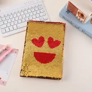 Close-up of a sparkling sequin notebook with heart eyes emoji design on white desk.