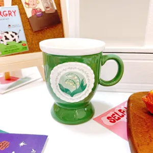 Green ceramic coffee mug with lily of the valley pattern and footed base for home office use.
