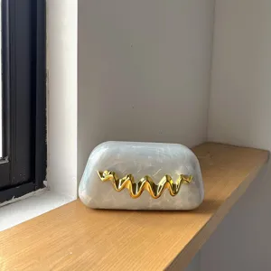 Detailaufnahme einer eleganten Acryl-Clutch mit Marmormuster und goldenem Metallwellenverschluss.