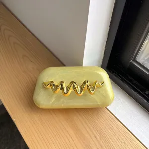 Detailansicht einer gelben Marmor-Acryl-Abendtasche mit goldenem Wellen-Metallverschluss.
