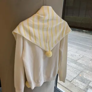 Detail shot of white knitted sweater featuring yellow striped collar and cute pom pom decorations.