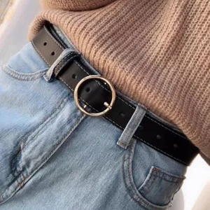 Detail shot of a black leather belt with a gold round buckle and white stitching worn on blue jeans.