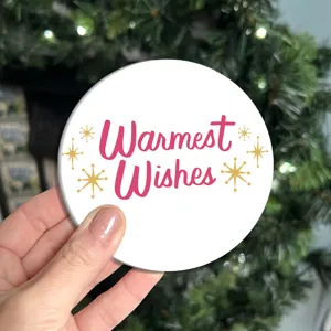Gros plan d'un sous-verre de Noël rond blanc avec le texte Warmest Wishes et des étoiles.