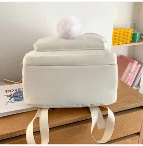 Detail view of white canvas backpack with fluffy pom-pom charm on top, daily casual campus style.