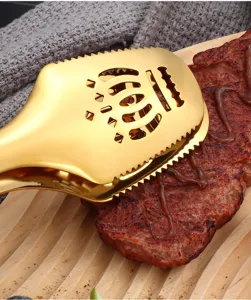 Gold stainless steel kitchen food tong detail, crown cutout design for steak and barbecue use.