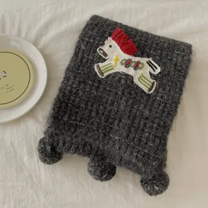 Detailed view of dark gray knitted winter scarf with cute little horse embroidery and fluffy pom poms.
