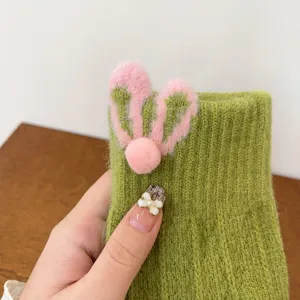 Dettaglio in primo piano di guanti lavorati a maglia verde con orecchie e pon pon rosa.