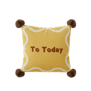 Yellow throw pillow with brown pom poms, white wavy border and To Today text for sofa bed.