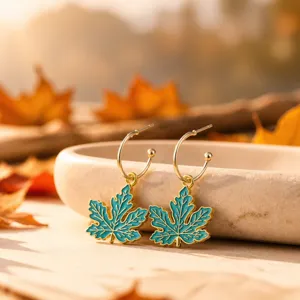 Close-up detail shot of the golden hoop earrings featuring vibrant teal enamel maple leaf pendants.