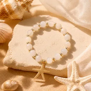 Detailaufnahme eines weißen Perlenarmbands mit goldenem Seestern-Anhänger auf Sand.