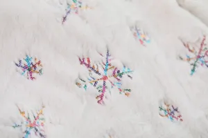 Close-up of white plush Christmas tree skirt with colorful sequin snowflake embroidery detail.